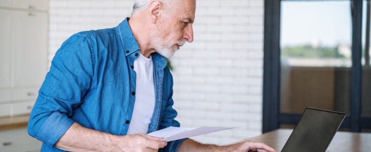 Serious middle aged man with paper document at home office checking financial taxes fees and reviewing bank account on laptop. Focused mature male in casual clothes paying medical insurance online.